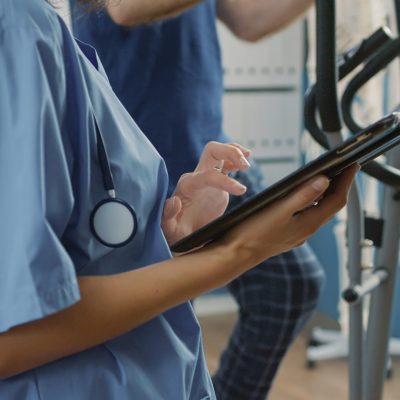 Health specialist in uniform using digital tablet to help senior patient with physical injury and mechanical disorders. Nurse holding device at muscle rehabilitation procedure. Close up.